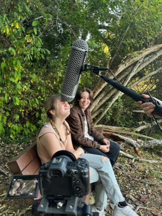 Two actors on a bench during filming, with a boom microphone and camera in frame