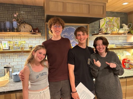 Four cast and crew members in a kitchen, one holding a script